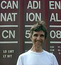 Head and shoulders photograph of a bearded smiling man in white T-shirt standing in front of a freight car bearing the words "Canadien National" and loading information