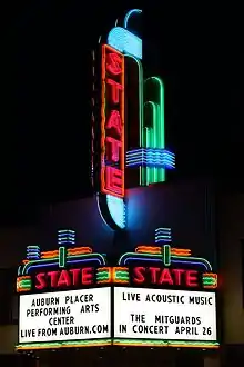 Photograph of a large, elaborate neon sign at night. The word "STATE" is written vertically in red neon tubing on a tower above a marquee. The marquee sign proper below the tower also has an elaborate neon tubing design, including the word "STATE" written horizontally in red neon tubing above each of the two panels facing the camera. A reader board on the front-facing panel has black lettering that says "AUBURN PLACER/PERFORMING ARTS/CENTER/LIVE FROM AUBURN.COM". A second reader board on a side panel says "LIVE ACOUSTIC MUSIC//THE MITGARDS/IN CONCERT APRIL 26".