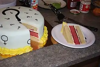 A cake frosted in white with black question marks and Mars and Venus symbols; the cake has been cut open and a piece sits on its side on a paper plate, revealing a pinkish middle layer.