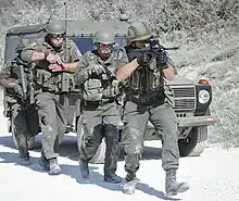  Four soldiers in green military attire walk in a line in front of a green military vehicle holding guns.