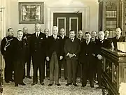 A sepia photo of fourteen men in suits standing in two rows