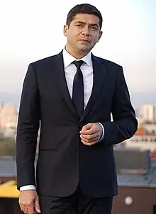 dark-haired white man wearing black suit and white shirt, standing, looking at camera