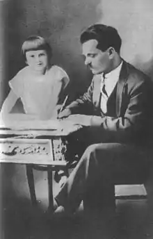 Photograph of Nestor Makhno writing at his desk, while his young daughter Elena climbs over the desk