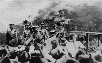 People gathering around a man standing on what is possibly a podium, reading a declaration, all of them wearing army- or related uniforms