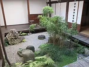 A garden courtyard with an en about 30 centimetres (12 in) above the pale gravel, feature stones, a rain chain, and a planting of bamboo, ferns, grasses, and a creeping plant with small round leaves.