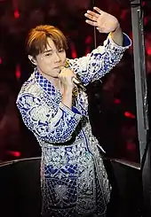 Close-up shot of Hins Cheung holding a microphone in his right hand and waving to the audience with his left hand