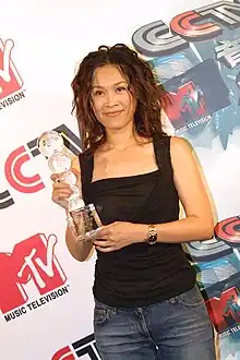 East Asian woman wearing blue jeans and a black top, standing on a podium, holding a trophy, with a smile on her face