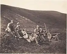 A group of hunters sitting outside, all wearing tweed clothing and carrying long guns.