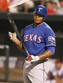 baseball player in a baseball uniform with a blue jersey that says "TEXAS" in white letters. He is holding a bat in his left hand and wearing a blue batting helmet.