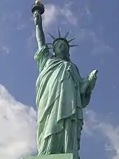 The Statue of Liberty as seen from below. The green statue is a female figure wearing a toga and crowd, while holding a torch above in her right hand. There is a blue sky and white clouds behind it.