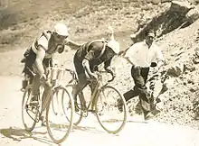 Two cyclists on bicycles, one cyclist with his hand on the back of the other cyclist. In the background, one man is running in the same direction.