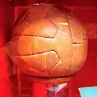 12-panel brown leather football on a plinth of hardwood, with a brass panel inscribed with the names of the victorious team members, photographed through the glass of a display case