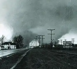 Two greyish tornado funnels silhouetted against a pale sky on either side of a road behind telephone poles