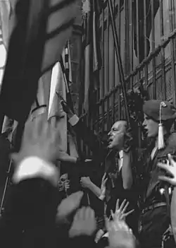 A man giving a fascist salute to a crowd, next to him stands a young man wear military style clothes and a beret