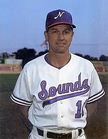 A man in a white baseball jersey with "Sounds" written on the front in red and blue and a blue cap with a white "N" on the front smiling with his hands behind his back.