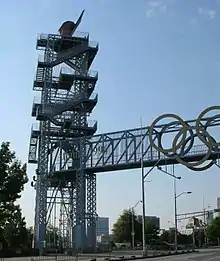 Complex structure of crosspieces apparently about three or four stories high in silhouette, with bridge like appendage bearing Olympic rings at second floor