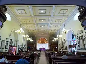 A symmetrical photo looking down the nave towards the apse and chancel showing the corners of the underside of the western gallery at its edges and the pews with some people sitting in them