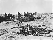  An artillery gun and its crew in a desert. A pile of shell cases is in the foreground.