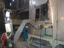 A robot with a metal arm is using its grinding wheel to cut into a wall, sending a shower of sparks upward.