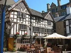 A timber-framed building, three floors high with a gabled roof. There are tables and chairs outside