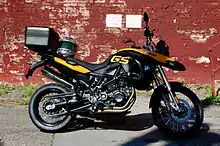 Black and yellow BMW F800GS with topbox and motorcycle helmet on the seat, parked in front of a brick wall covered in peeling red paint