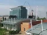 View from above of a building with white poles sticking up from its sides and the cables connecting them.  A taller building is in the background and a connected brick building is visible on its right.