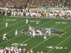 American football players in formation at the 50-yard line on a green field.