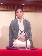 A Japanese man kneeling wearing a cream kimono and a blue jacket.