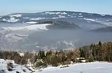 A valley in low, partially forested mountains seen in wintertime, covered with snow. At the bottom is a village, almost obscured by a layer of grayish-brown air