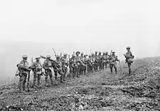 A line of soldiers in battle equipment face another soldier who is addressing them on a gentle slope.  Behind them, smoke or fog obscures the rest of the terrain.