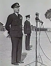 Man in dark military uniform with peaked cap in front of microphone