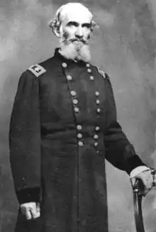 Black and white photo shows a white-haired man with a beard. He wears the dark military uniform of a one-star general and holds a cane in his left hand.