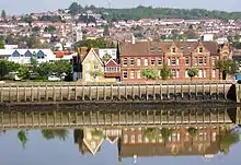 A river scene with wharf and above it a two-storey red brick building. The building has decorative brickwork and plenty of windows. Along the facade are two three-storey decorated gable features.