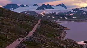 A road through a barren landscape with a lake to the right and mountains in the background