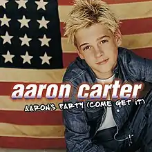 An image of a teenage boy with blond hair, wearing a white t-shirt, jean jacket and jeans, with the United States flag behind him.