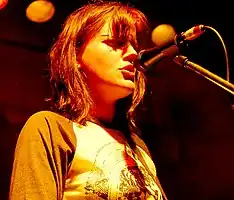 May is singing into a microphone on its stand. Her should length hair is brown and the fringe covers her eyes. She stares forward, wearing a brown sleeved tee-shirt with a design printed on front. Stage lights are visible behind and above her.