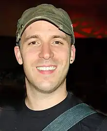 Adam Kowalczyk wearing olive green cap, black t-shirt, with strap over left shoulder and small earring in left ear, grinning at camera