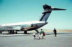 Rear three-quarter angle of twin-engined passenger jet with high tail plane, parked on tarmac with four people walking towards it