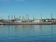 Adelaide tied up alongside at a wharf. The majority of her communications and radar masts have been removed, she carries no weapons, and several large squares have been cut into the ship's hull.