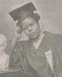 A young African-American woman wearing a mortar board cap and a blouse with a high lacy neckline. There is a ribbon pinned to her academic robes. She is wearing glasses, and resting her had on her right hand.