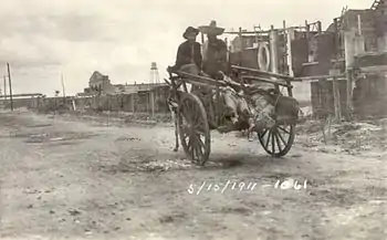 Two men in sombreros riding in a donkey-cart with a line of feet sticking out the back. They are riding down a dirt street away from the camera, with a line of buildings on the right.  Dated 15 May 1911.