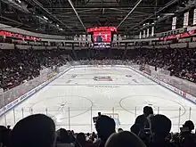 A full hockey arena with a clean ice surface. The arena is full of people. We are looking straight ahead from the home bench.