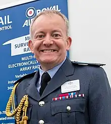 Portrait photograph of head and chest of Sir Richard Knighton, taken indoors in front of a display banner.  Knighton is wearing his Royal Air Force 'Number 1' service dress without head-dress, with medal ribbons, but without actual medals, and above the ribbons is his name badge.  Knighton has a broad smile, clean-shaven, and brushed-back light-coloured hair.