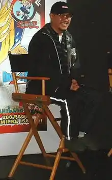 Akira Toriyama sitting on a chair smiling wearing black clothing with glasses and chin facial hair