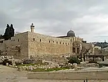 A photograph showing the corner of a high stone wall with several openings at the top, above which rise a minaret and a gray dome