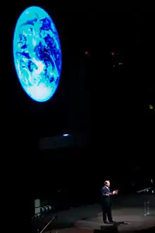 A man is standing on a stage in front of a giant projection of the Earth