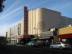 A color photograph of a movie theater facade and street scene. The theater is designed in the Streamline Moderne style, with Moorish elements, and a blade which reads "Alameda" standing vertically above a marquee announcing films.