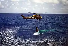 A green helicopter hovers low over the water, with the Mercury capsule suspended below. The helicopter has "Marines" written on it, and the number "44"