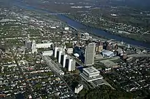 A developed city center with a complex of tall modern white towers in the middle and a river running across the upper right corner. Older buildings are above and to the right of the modern ones; beyond them are areas of just houses amid green trees