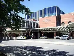 Three-story brick building with blue windows; a glass pedestrian bridge travels from building to unseen parking garage on left, crossing entrance road. "ALBANY INTERNATIONAL AIRPORT" sign is visible on side.